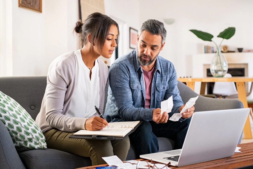 Casal sentado num sofá a analisar vários recibos e a consultar o computador portátil que está em cima da mesa à sua frente.