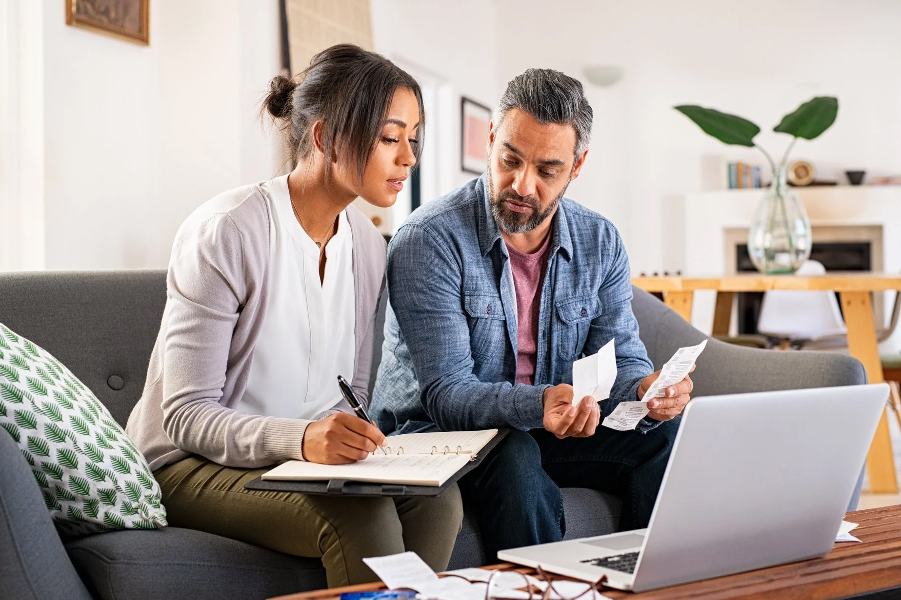 Casal sentado num sofá a analisar vários recibos e a consultar o computador portátil que está em cima da mesa à sua frente.