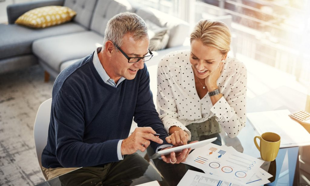 Casal de meia idade sentados a analisar um tablet e vários gráfico em vários papéis.