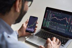Homem sentado a consultar uma aplicação financeira no smartphone e no computador portátil.