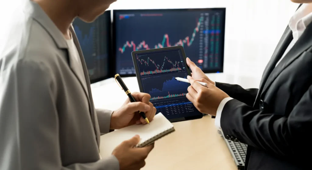 Computador e tablet com gráficos de bolsa. Homem e mulher a conversar e fazer apontamentos.