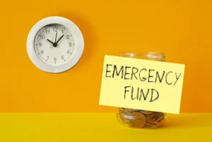 Um relógio de parede e um post-it com a nota "Emergency Fund" colado num frasco cheio de moedas.